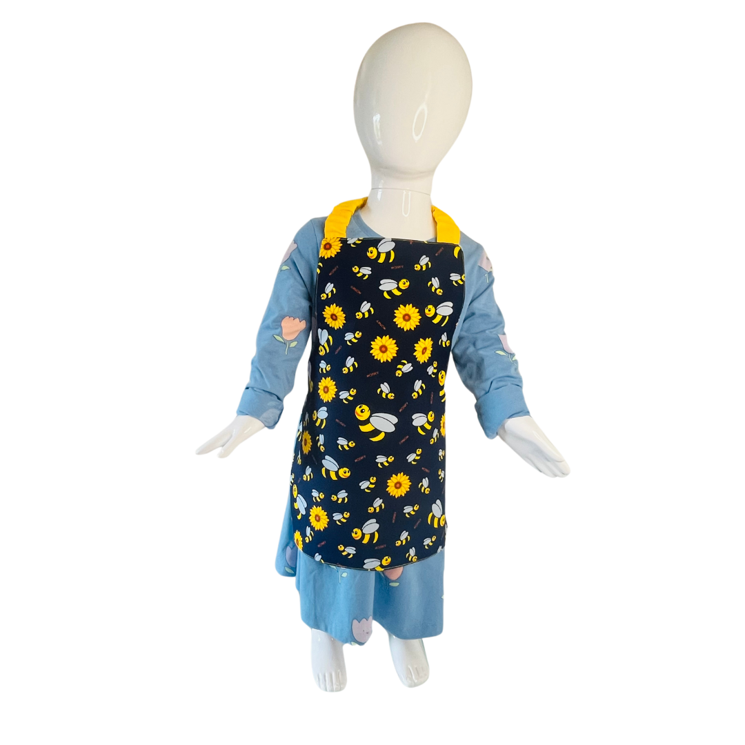 A child’s apron and matching oven mitt set. Pattern is navy and yellow with cartoon bumble bees and yellow flowers all over. Styled on mannequin.