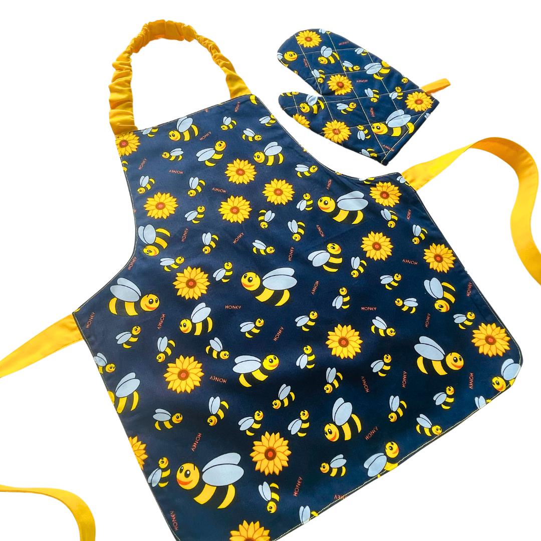 A child’s apron and matching oven mitt set. Pattern is navy and yellow with cartoon bumble bees and yellow flowers all over.