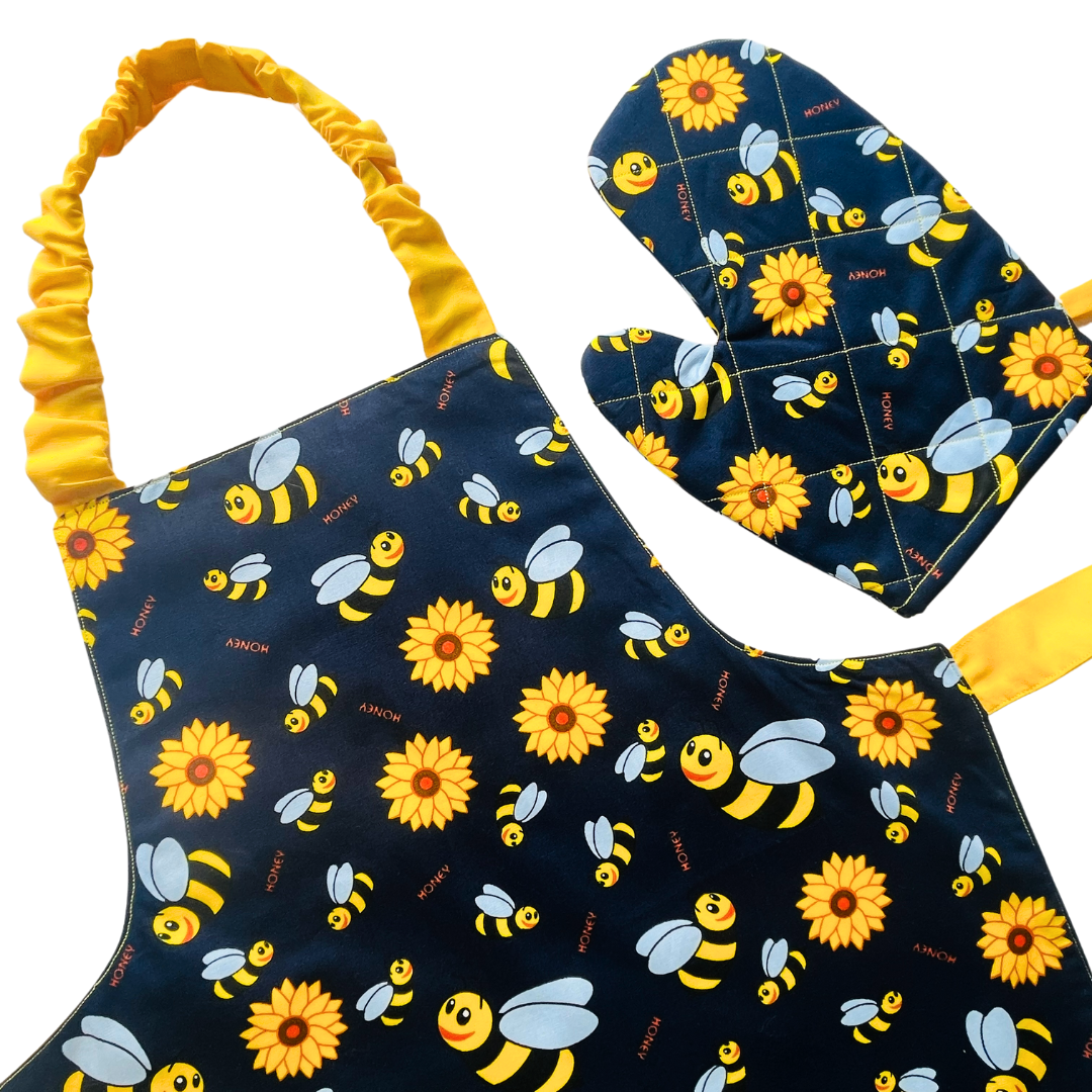 A child’s apron and matching oven mitt set. Pattern is navy and yellow with cartoon bumble bees and yellow flowers all over.