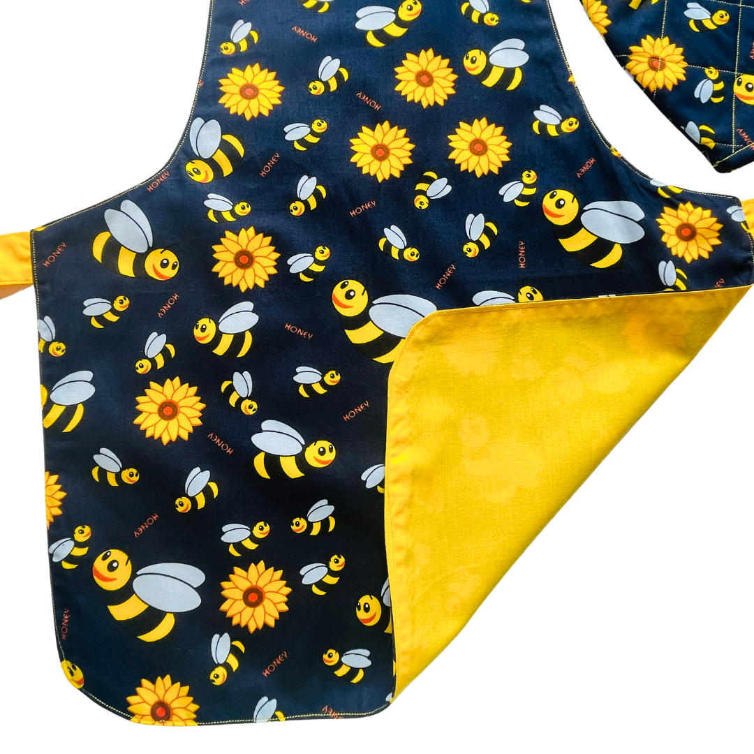 A child’s apron and matching oven mitt set. Pattern is navy and yellow with cartoon bumble bees and yellow flowers all over.